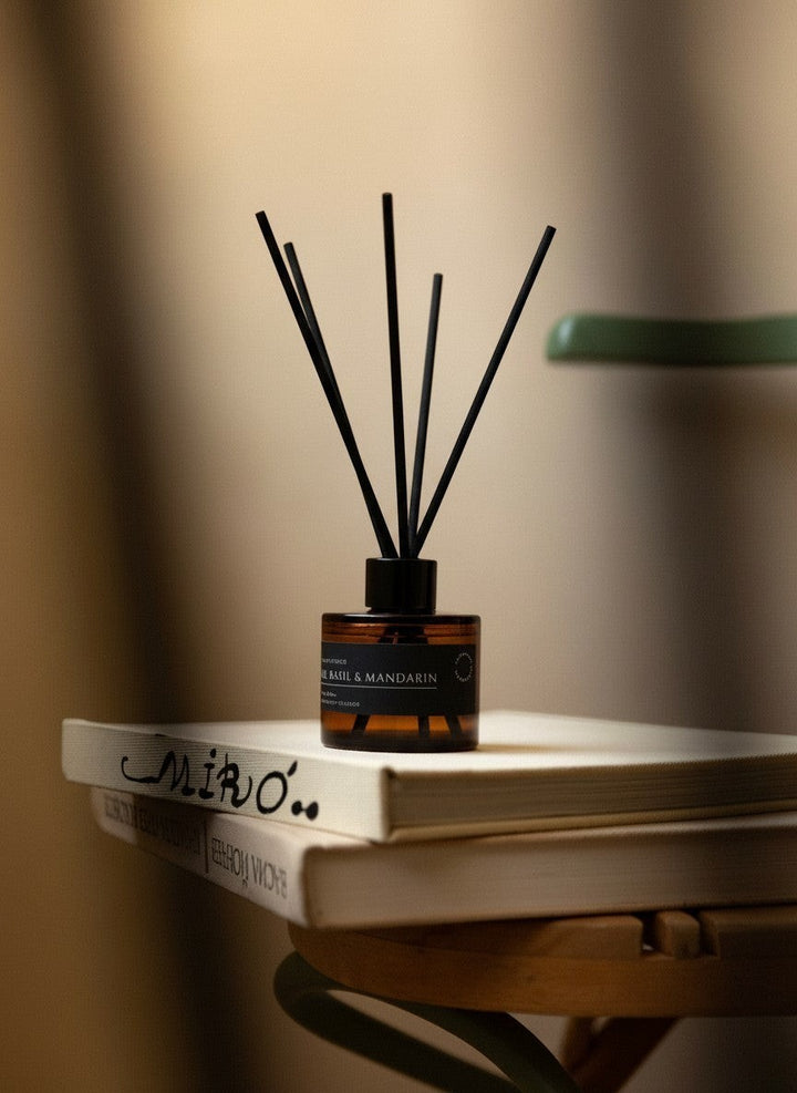 Glim & Glam Lime, Basil & Mandarin reed diffuser in an amber glass bottle with black reeds resting on stacked books.