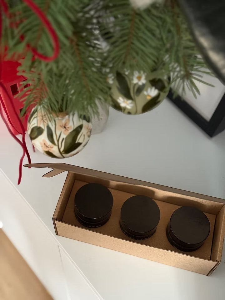 Glim & Glam Slow Season gift set of three mini soy candles in black jars, nestled in a cardboard box, with festive ornaments and pine branches above.